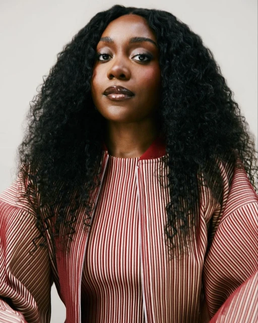 Portrait of a dark-skinned woman with curly black hair, wearing a striped jacket, set against a neutral background.