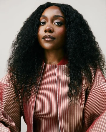 Portrait of a dark-skinned woman with curly black hair, wearing a striped jacket, set against a neutral background.
