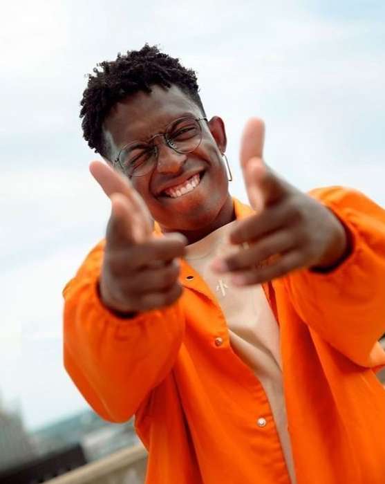 Smiling adult man poses with hand gestures wearing an orange jacket and glasses against an outdoor urban setting with a partly cloudy sky.