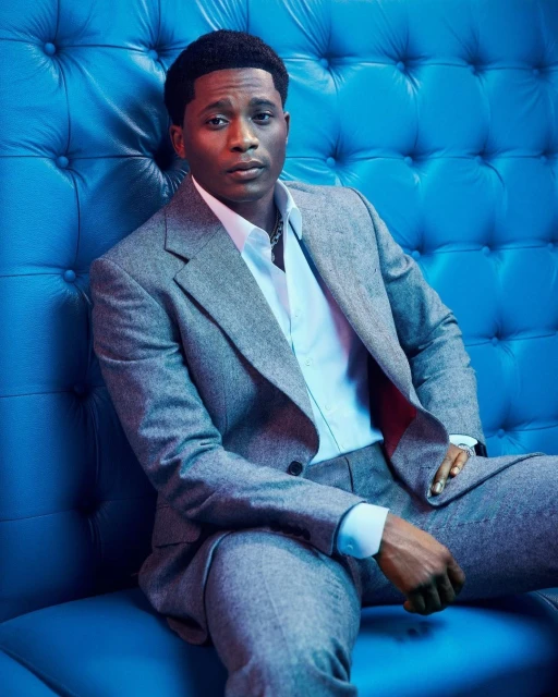 Portrait of an adult male in a gray suit and white shirt, sitting on a blue tufted surface under blue-toned studio lighting.