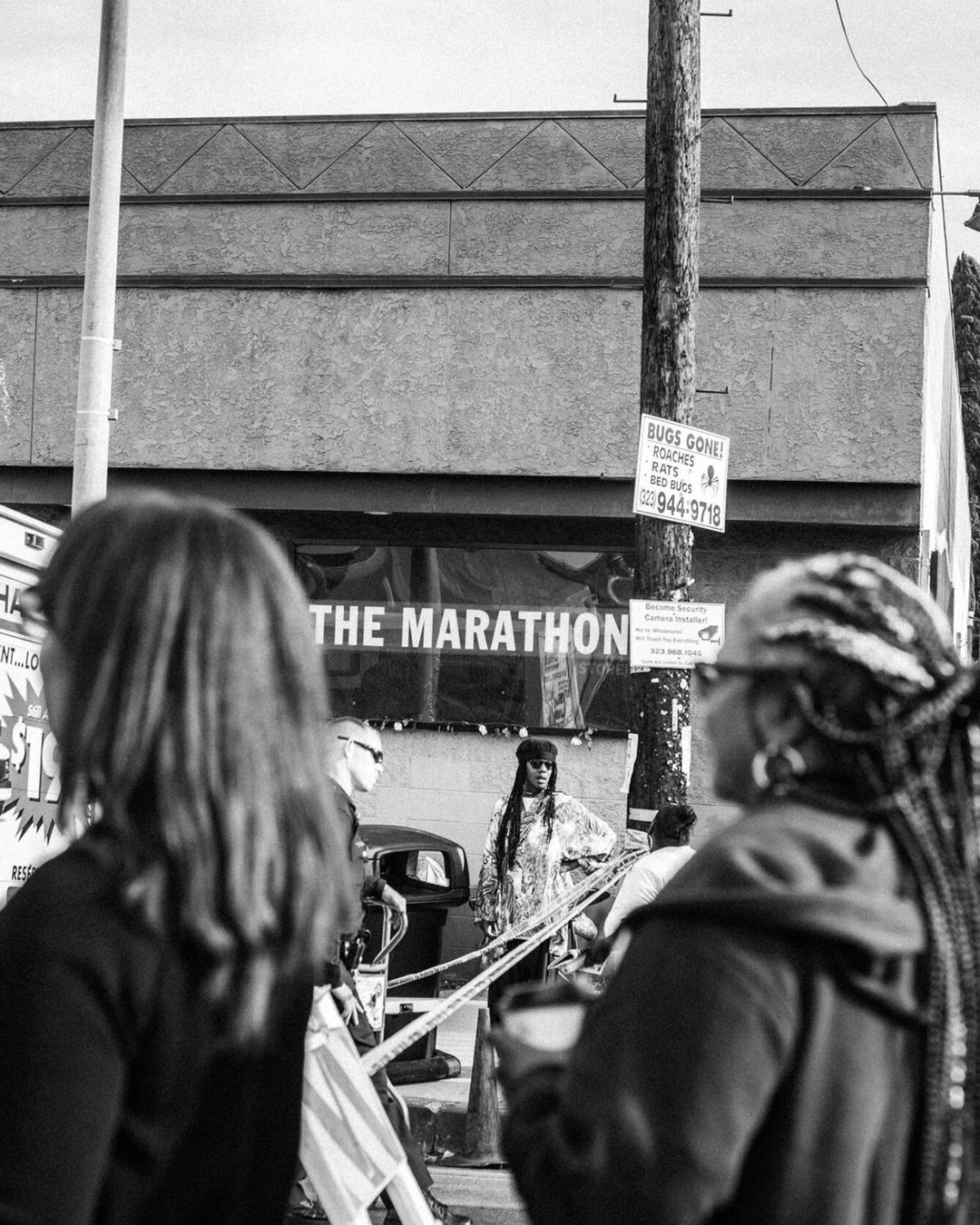 Black and white photograph of several people standing outside of a storefront with "The Marathon" sign, city street environment.