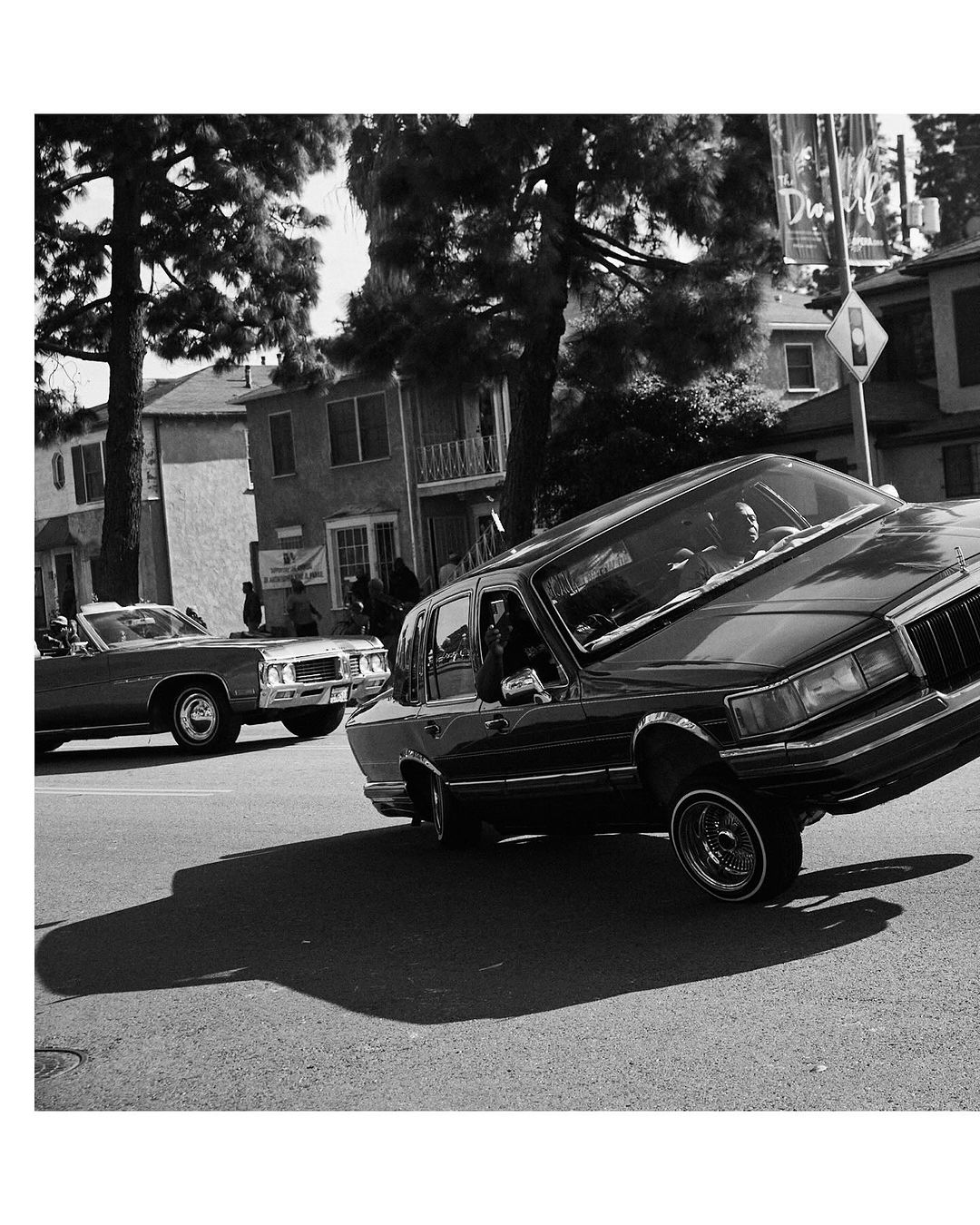 Monochromatic photograph of a classic lowrider car with a driver on a city street, showing car culture and urban environment.
