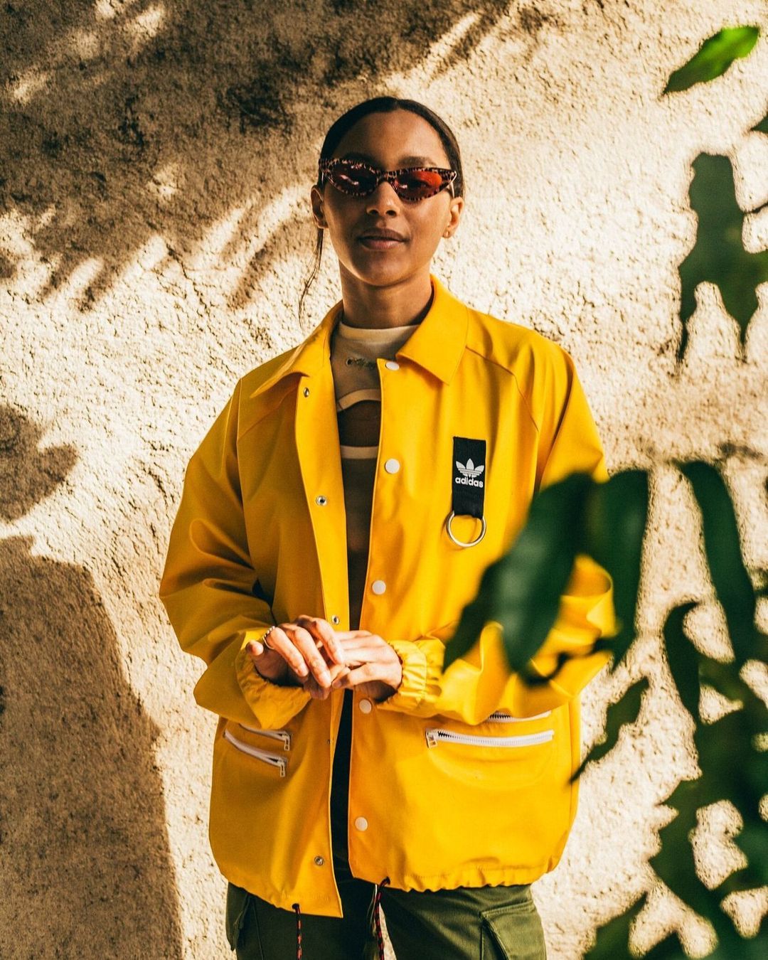 Portrait of an adult woman wearing a yellow Adidas jacket and sunglasses, posing outdoors in front of a textured wall with plants.