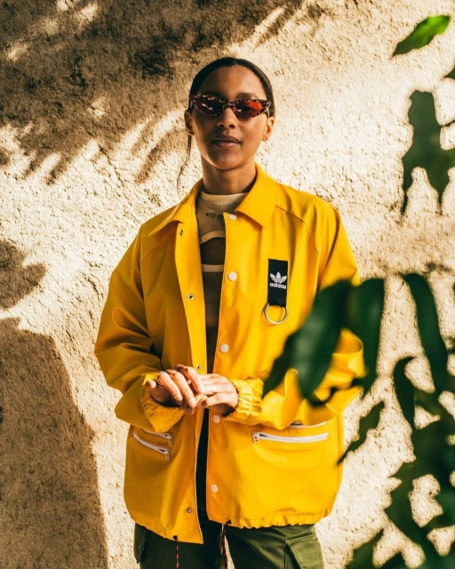 Portrait of an adult woman wearing a yellow Adidas jacket and sunglasses, posing outdoors in front of a textured wall with plants.