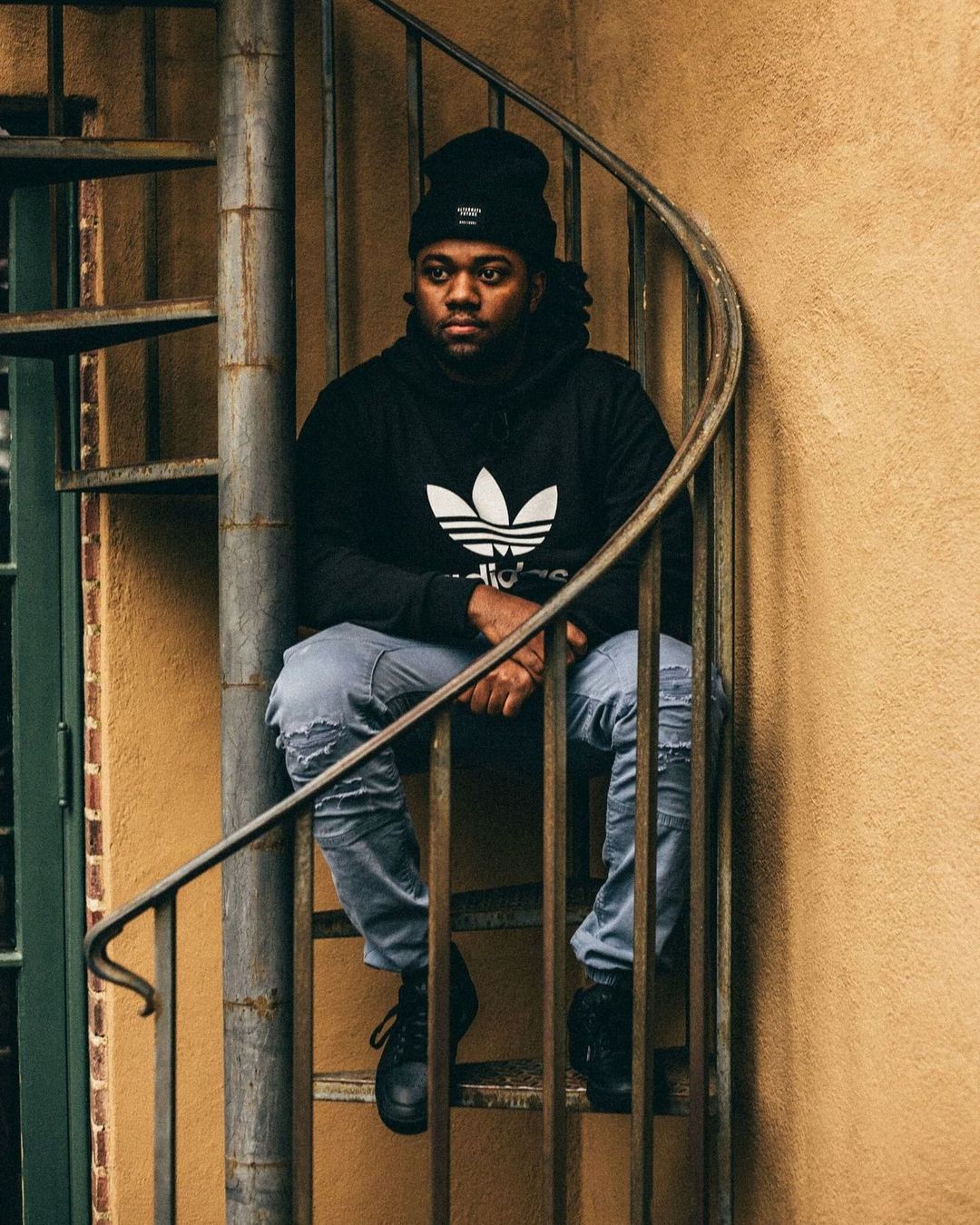 An adult male in a beanie and black Adidas hoodie sits on a spiral staircase outdoors, looking contemplative in a medium eye-level shot.