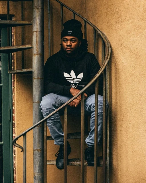 An adult male in a beanie and black Adidas hoodie sits on a spiral staircase outdoors, looking contemplative in a medium eye-level shot.