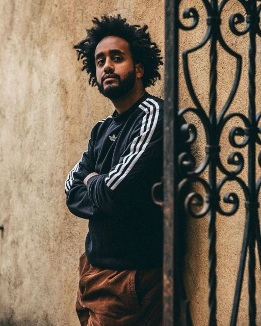 Portrait of an adult male with afro and beard, wearing a black Adidas sweatshirt, posing outdoors with arms crossed next to wrought iron gate.