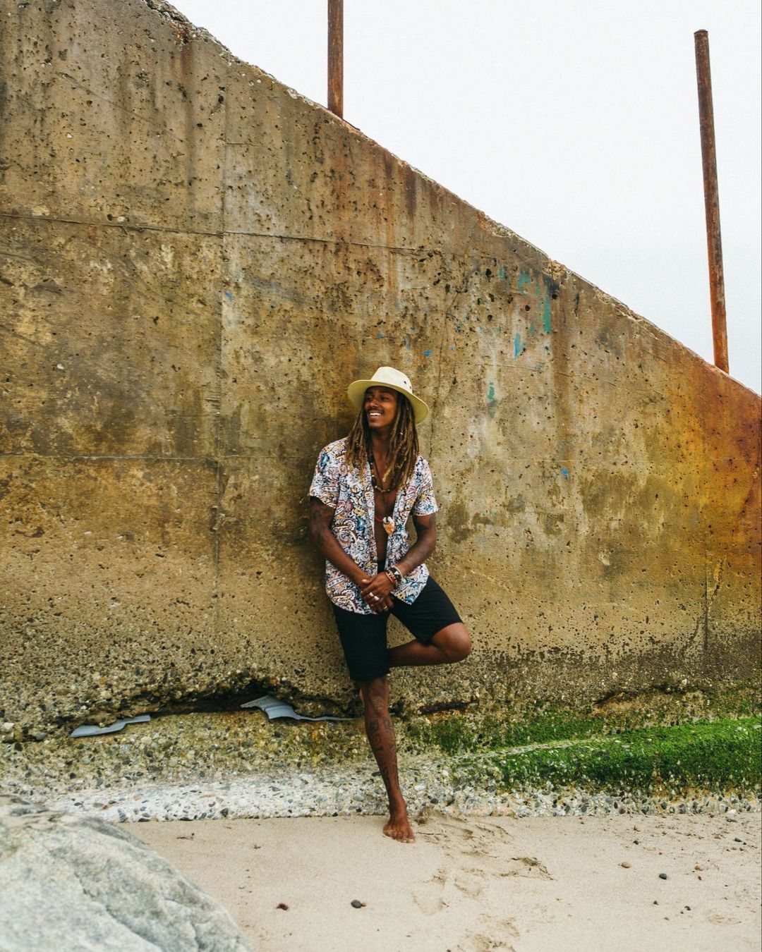 A man with long dreadlocks wearing a patterned shirt and straw hat leaning against a concrete wall at the beach.