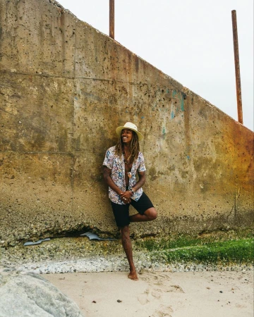 A man with long dreadlocks wearing a patterned shirt and straw hat leaning against a concrete wall at the beach.