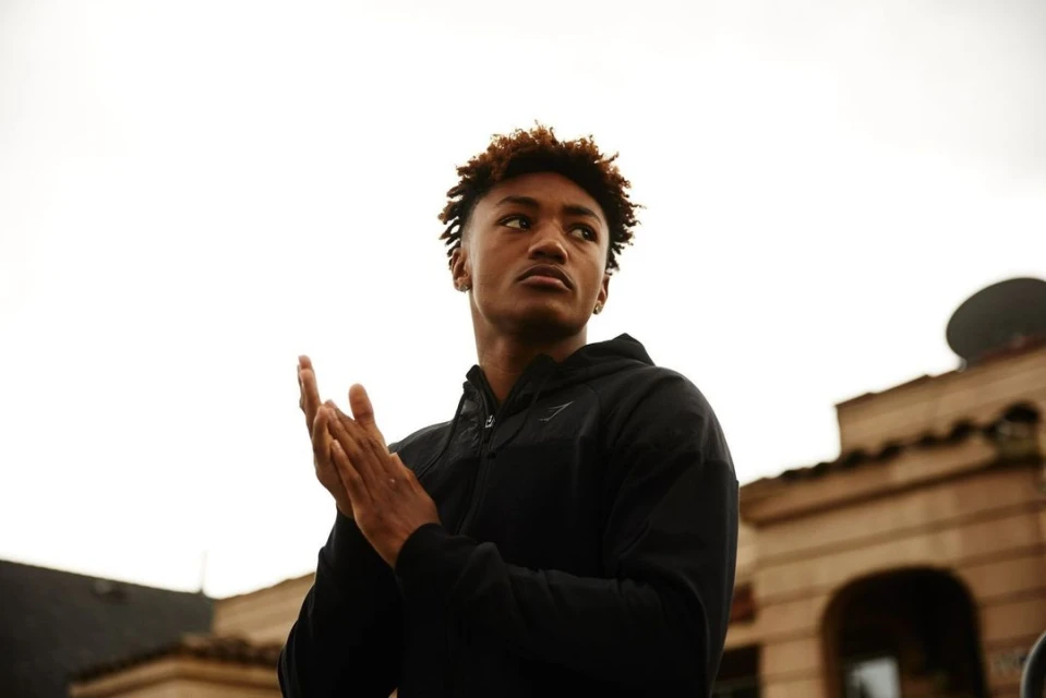 Low-angle shot of a young adult male with brown hair wearing a black hooded jacket, standing in an urban setting, looking upward.