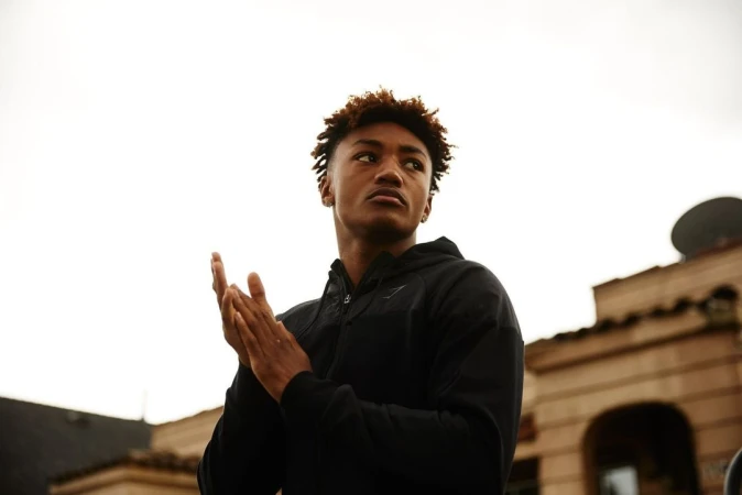 Low-angle shot of a young adult male with brown hair wearing a black hooded jacket, standing in an urban setting, looking upward.