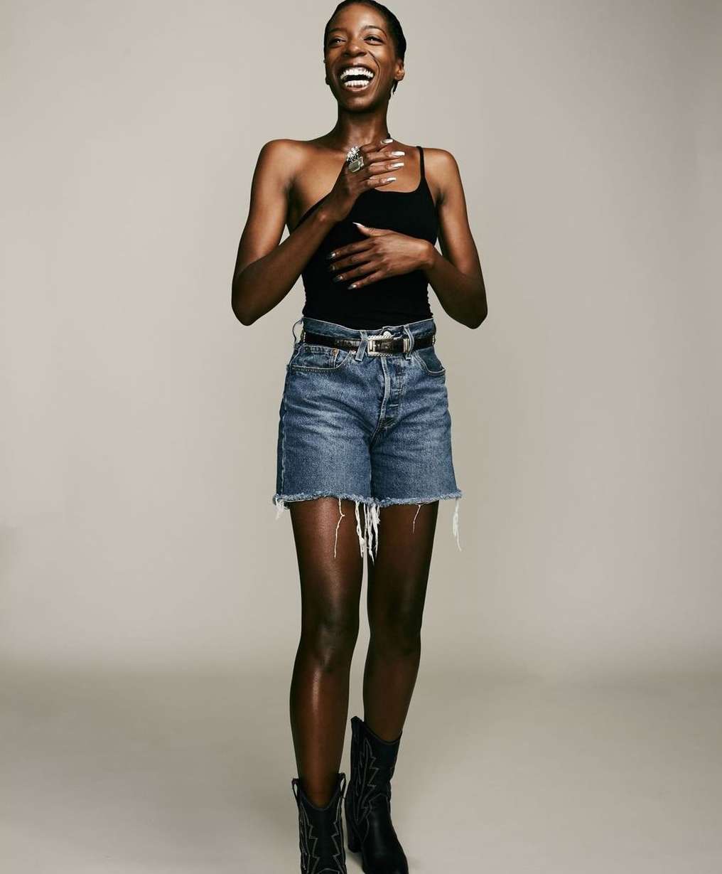 Smiling Black woman wearing a black top, denim shorts, and boots against a plain light grey background