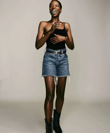 Smiling Black woman wearing a black top, denim shorts, and boots against a plain light grey background