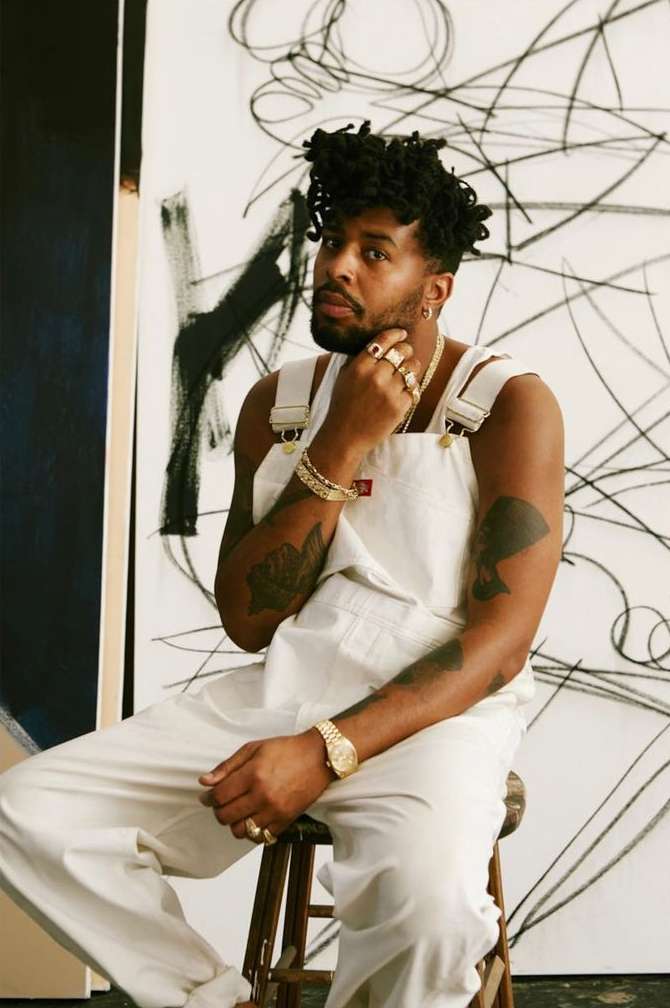 Portrait of a Black man sitting on a stool in white overalls and gold jewelry, with abstract art on canvas in the background.