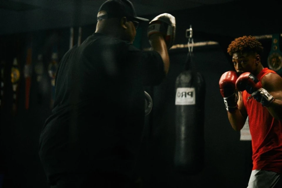 Two men boxing in a gym with a heavy bag, one with red boxing gloves and a red shirt, the other out of focus in a black shirt.