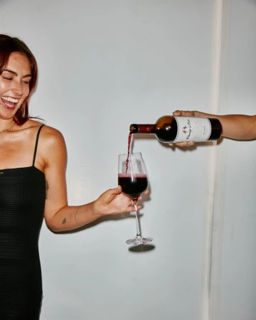 Smiling woman in a black dress holding a wine glass while red wine is poured from a bottle by another person