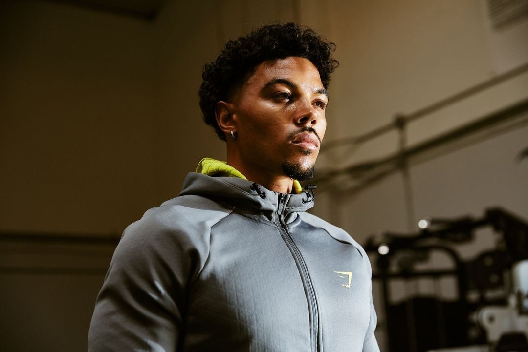 Athletic Black man in a gray hoodie looks to the side in an indoor setting, showcasing fitness wear and a focused gaze.