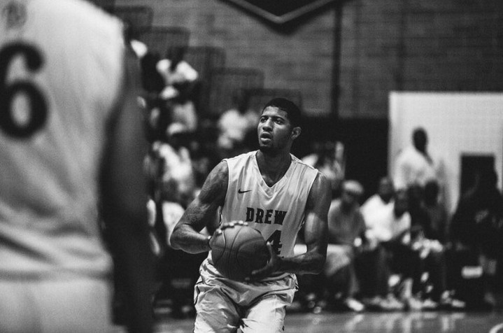 Monochrome action shot of a man in a tank top holding a basketball, poised to shoot, with a blurred crowd and gym background.