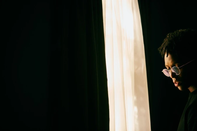 Portrait of a Black man wearing round glasses standing beside a bright window with sheer curtains.