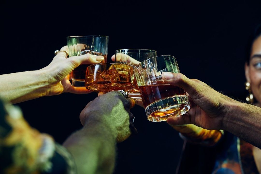 Four people toasting with glasses of amber liquid in a dimly lit indoor setting, celebrating with friends, in a casual gathering.