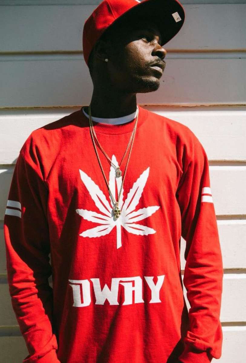 Adult male in red baseball cap and long-sleeve shirt with marijuana leaf design, gold chains, and a serious facial expression.
