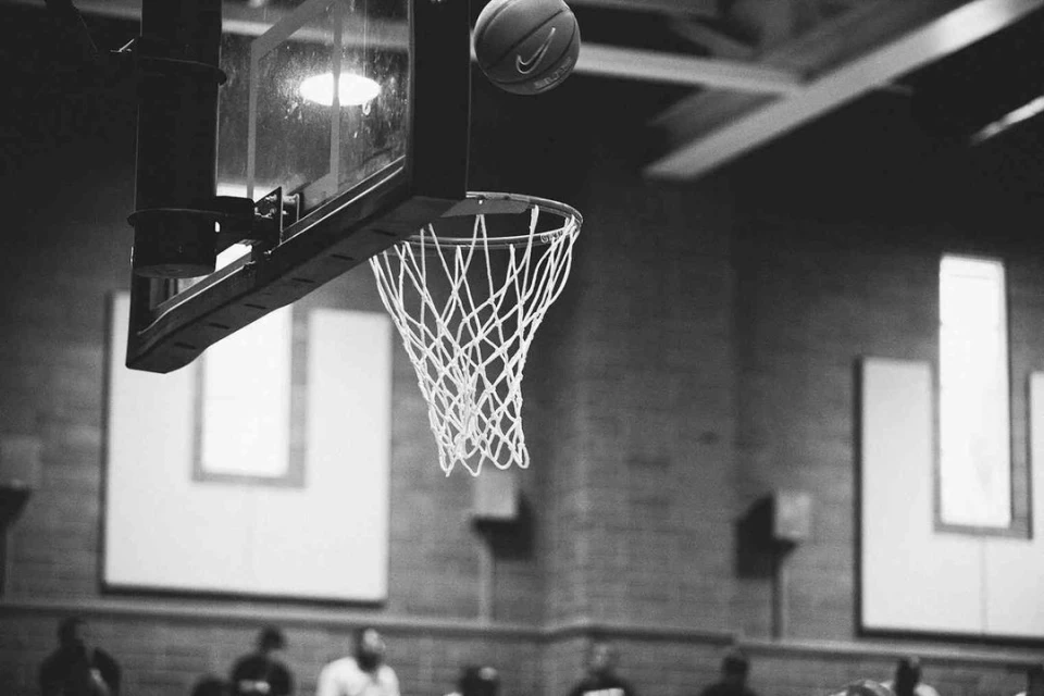 Monochrome image of a basketball going into a net attached to a backboard, people visible in the background in a gymnasium setting.