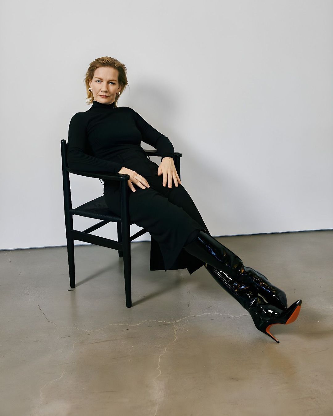 Elegant woman in black dress and shiny boots sitting in a chair against a neutral background, fashion studio shot.