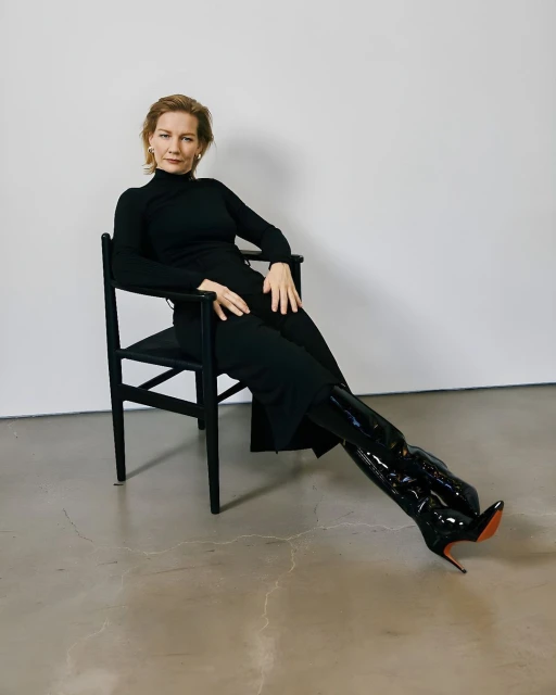Elegant woman in black dress and shiny boots sitting in a chair against a neutral background, fashion studio shot.