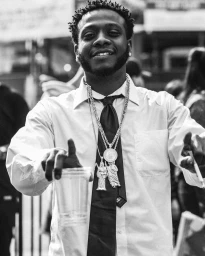 Black and white portrait of a smiling Black man wearing a white collared shirt, dark tie, and layered silver chains.