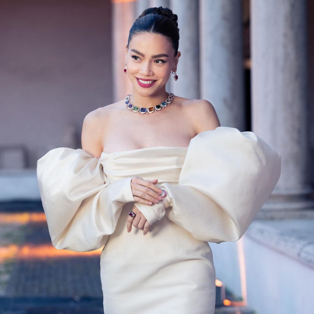 Elegant woman in an off-shoulder dress with statement sleeves and colourful jeweled necklace posing in front of an architectural backdrop.