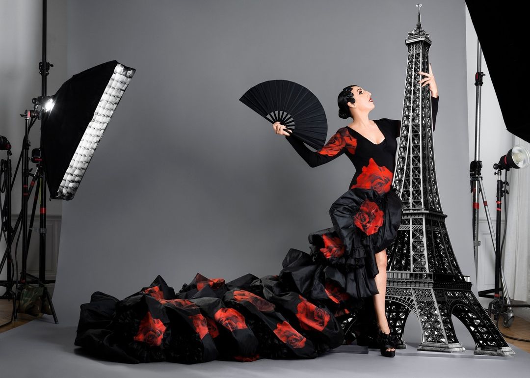 Woman in a black and red rose dress poses with a large Eiffel Tower prop in a photo studio, holding a fan, next to studio lighting.