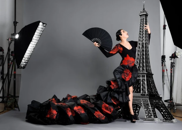Woman in a black and red rose dress poses with a large Eiffel Tower prop in a photo studio, holding a fan, next to studio lighting.