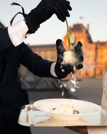 Elegant food presentation with gloved hands holding a white rose over a plate, set against a blurred cityscape backdrop at daytime.