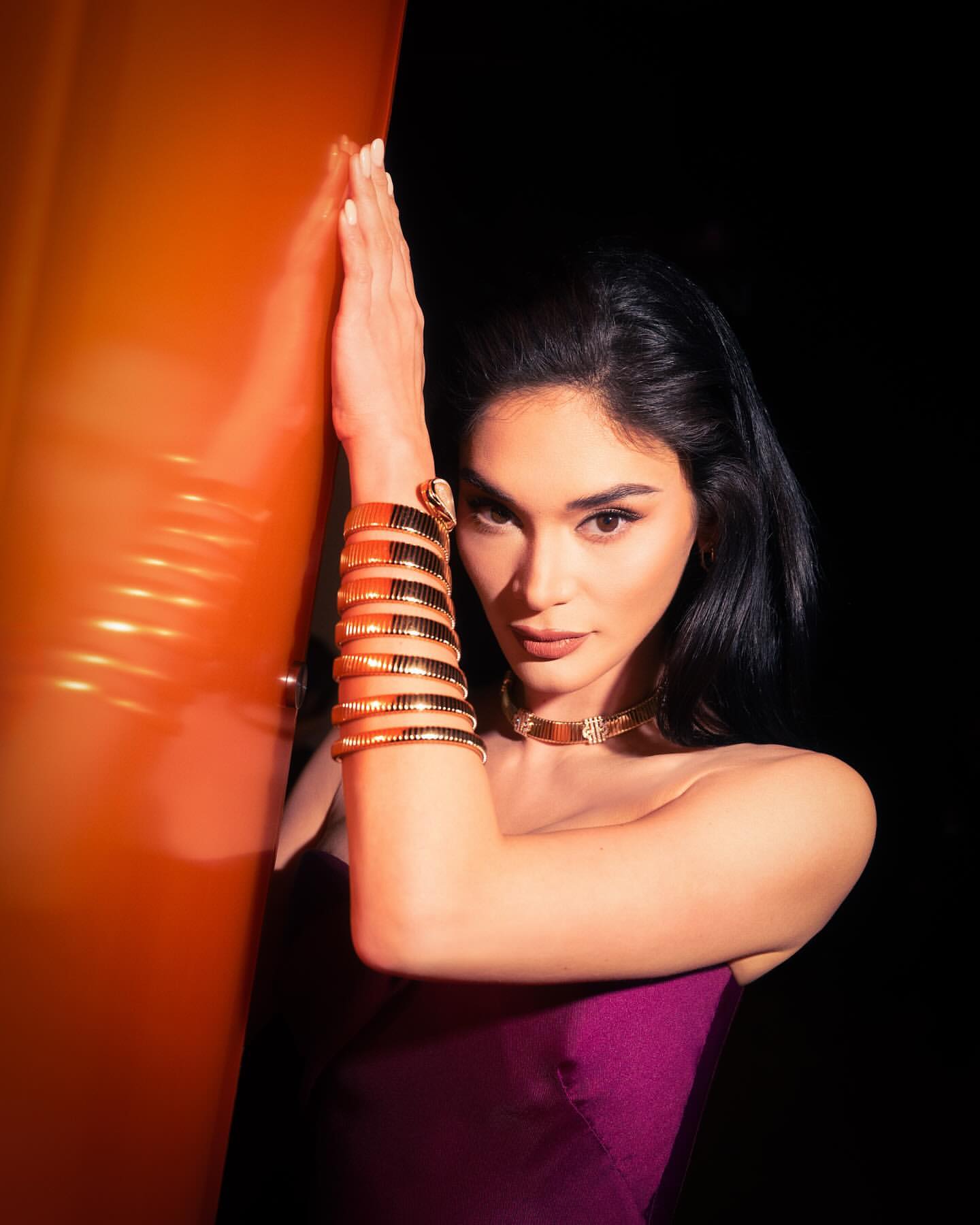 Brunette woman in a purple dress poses with gold jewelry and her arm resting on a orange structure in a dark indoor environment.