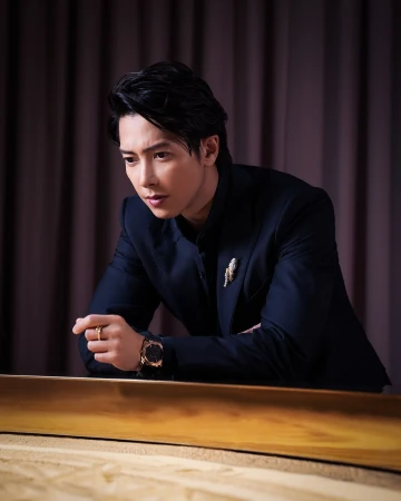 Headshot of a stylish Asian man in a dark suit with a gold brooch, leaning against a wooden surface, wearing a gold watch and ring.