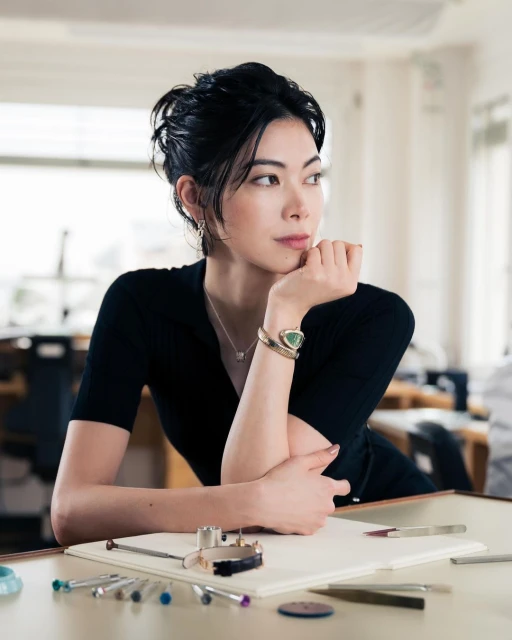 Adult woman sits at table in bright indoor setting, wearing black top and bracelets; tools for watchmaking are displayed on the table.