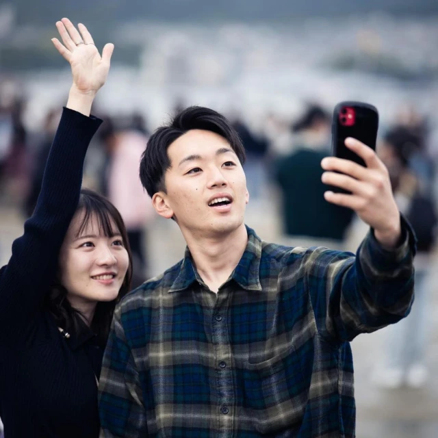 Smiling Asian couple takes a selfie outdoors; a young man holds the smartphone while the woman waves her hand with blurred background.