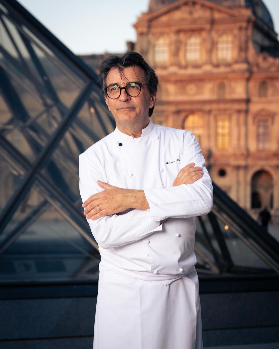 Elegant portrait of a chef in a white uniform with the Louvre Pyramid and Louvre Museum in the background on a sunny day.