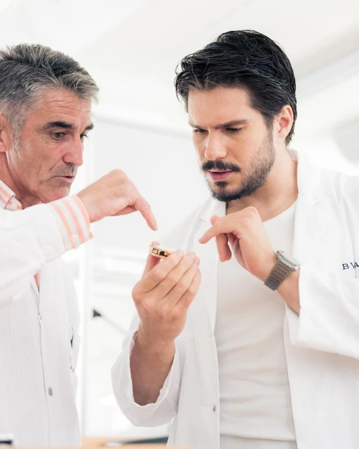Two adult men in lab coats examine a small, gold-colored object, indoors under bright lighting, focusing on the details of the object.