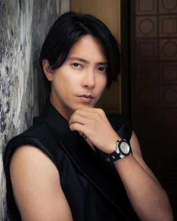 Portrait of an Asian man in a sleeveless black vest leaning against a patterned wall, looking at the camera with a serious expression.