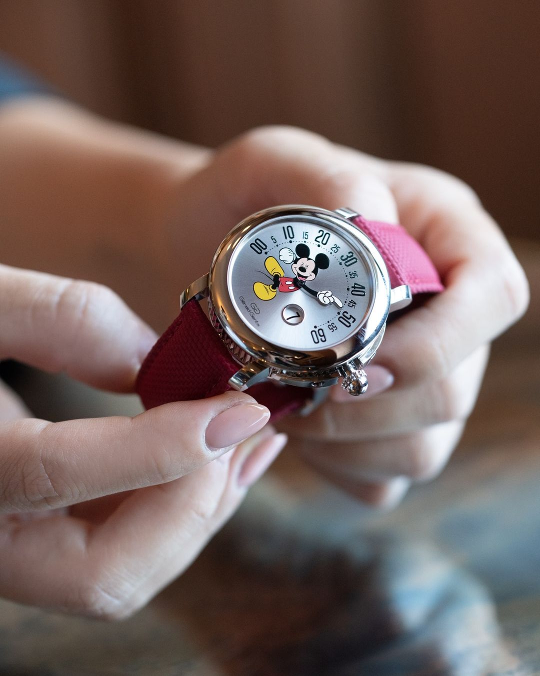 Close-up of a woman's hands holding a wristwatch with a silver face and red band, featuring a Mickey Mouse cartoon character.