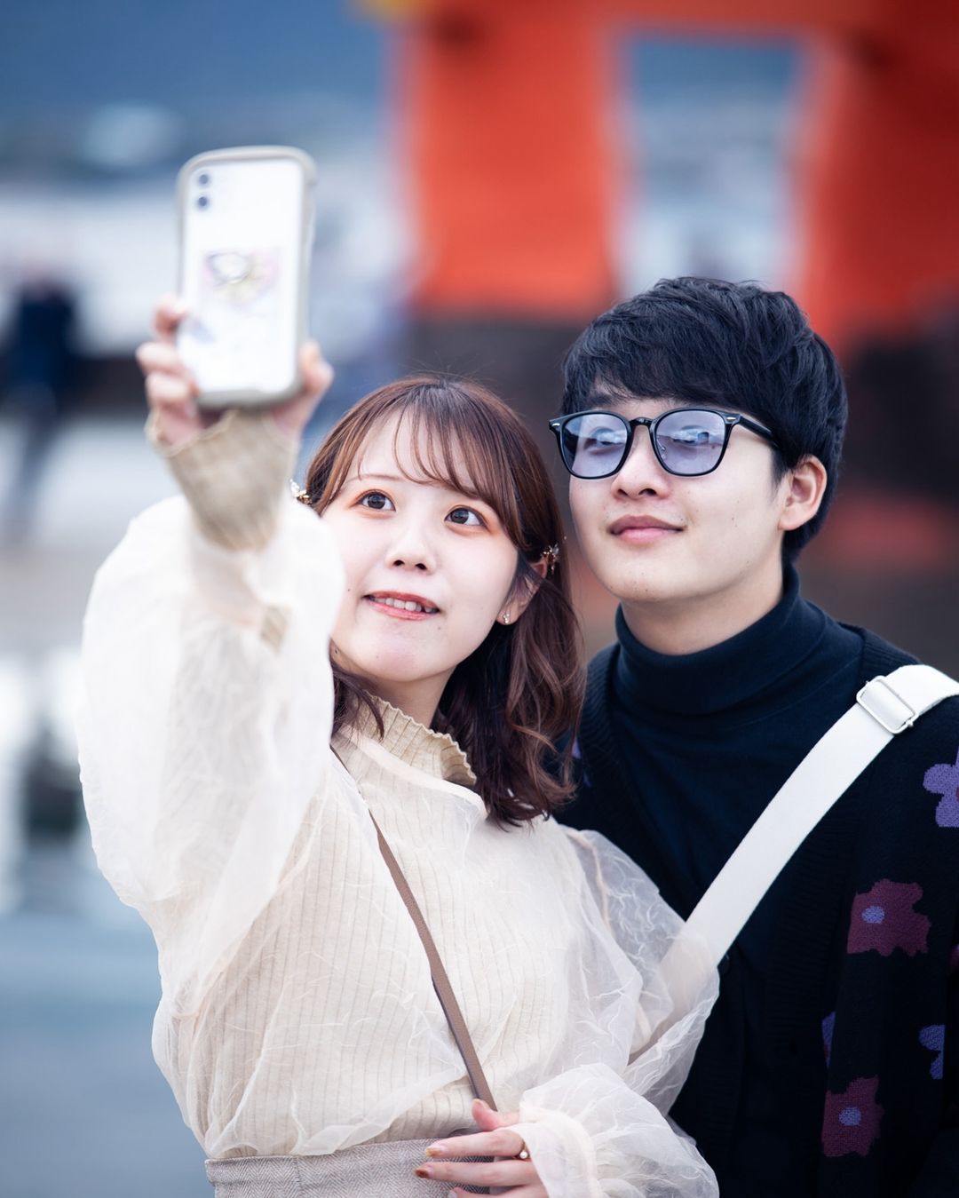 Young Asian couple takes a selfie outdoors in front of a red structure. The woman holds the phone, both smile, and wear casual outfits.