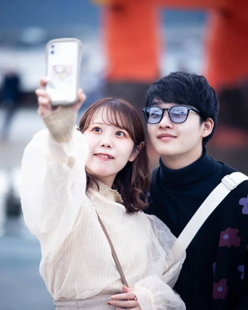 Young Asian couple takes a selfie outdoors in front of a red structure. The woman holds the phone, both smile, and wear casual outfits.