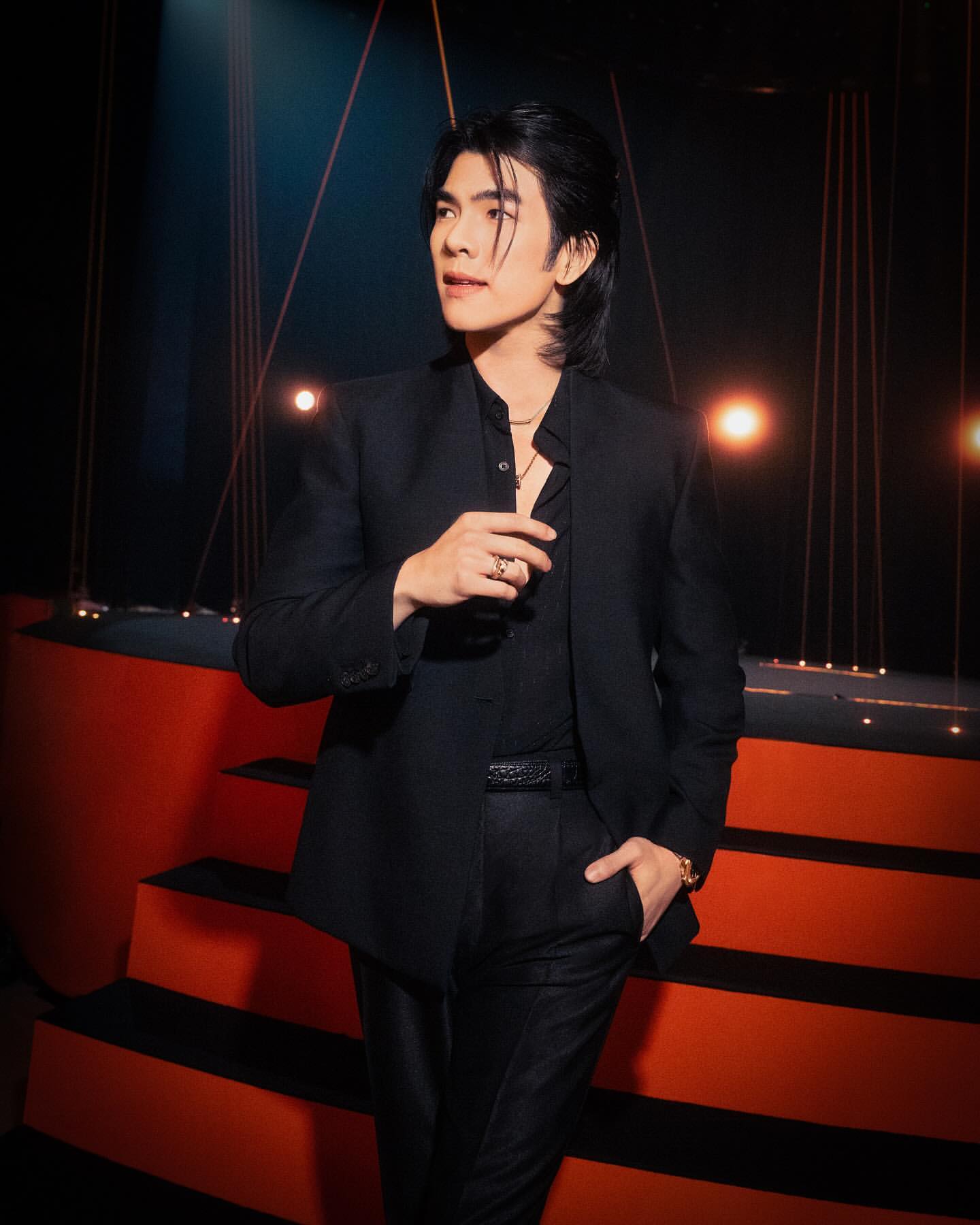 Portrait of a young adult male posing in a black suit against a dark backdrop with warm orange light, looking off to the left.