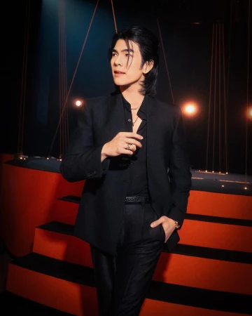 Portrait of a young adult male posing in a black suit against a dark backdrop with warm orange light, looking off to the left.