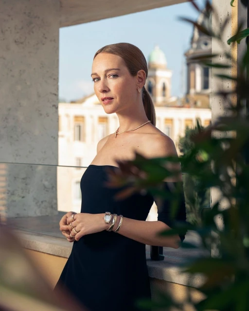 Woman in black dress poses on balcony overlooking Rome, Italy. She wears jewelry and has a neutral expression in this medium shot.