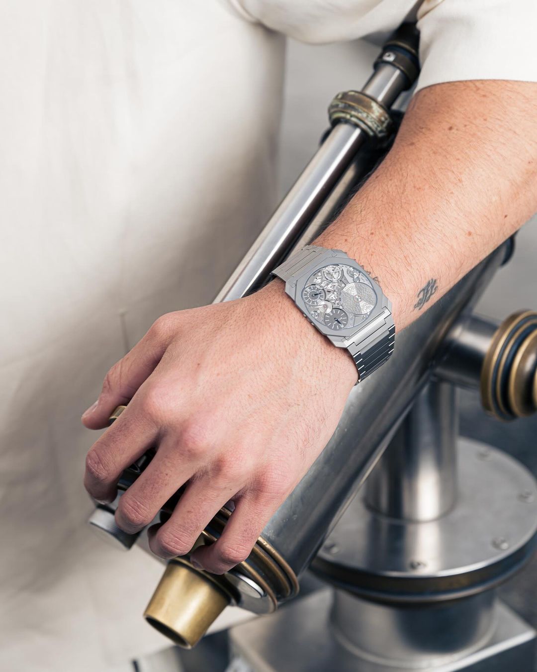 Close-up shot of a silver wristwatch on a man's wrist, posed with an industrial-style metal object in an indoor setting with natural lighting.