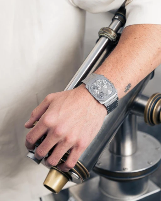 Close-up shot of a silver wristwatch on a man's wrist, posed with an industrial-style metal object in an indoor setting with natural lighting.