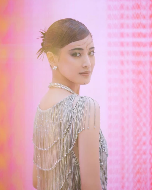 Portrait of an adult woman with dark hair and light skin wearing a pearl necklace and a silver fringed dress, looking over her shoulder.