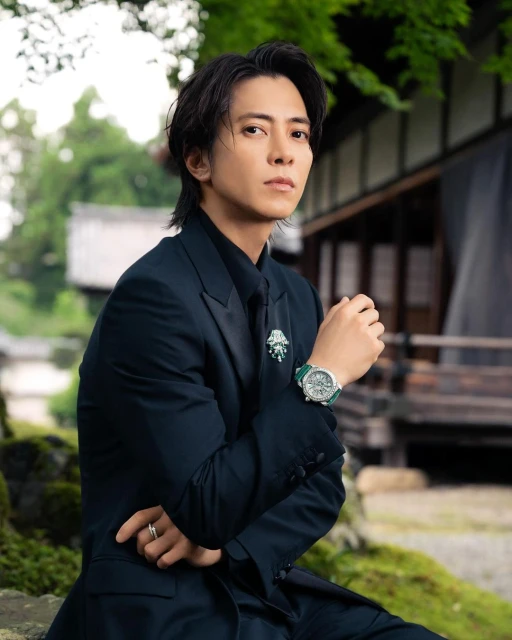 Portrait of an Asian man in a dark suit with a green watch sitting outdoors against a backdrop of traditional Japanese architecture.